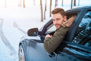 A man leans out of the driver side window of a vehicle while driving in the snow