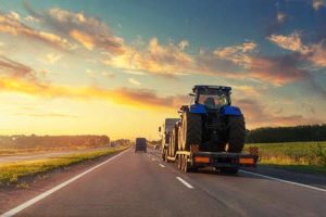 A semi carries a tractor on its trailer as it drives down a country road at dusk.