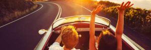 A man and woman ride in a vintage sports car on an open road