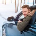 A man leans out of the driver side window of a vehicle while driving in the snow