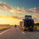 A semi carries a tractor on its trailer as it drives down a country road at dusk.