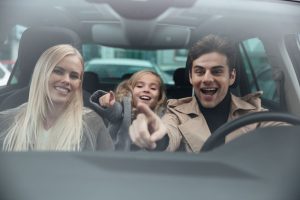 Photo of cheerful young man sitting in car with his wife and daughter. Looking aside and pointing.